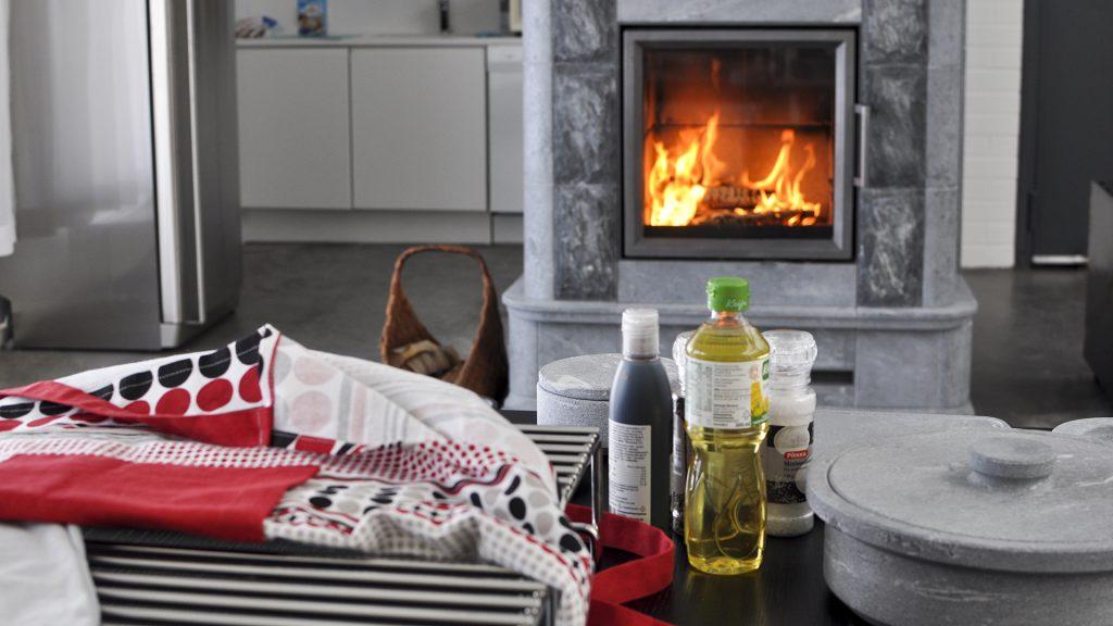 Ett mysigt vardagsrum med en braskamin i sten som eldas med ved, en korg vid sidan av och ett kök i bakgrunden. I förgrunden finns matlagningsingredienser, vikta textilier och köksredskap i sten på ett mörkt bord.