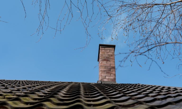 En skorsten i rött tegel avger svag rök ovanför ett vågigt tak med mörka tegelpannor, med nakna trädgrenar som ramar in den klarblå himlen i bakgrunden.