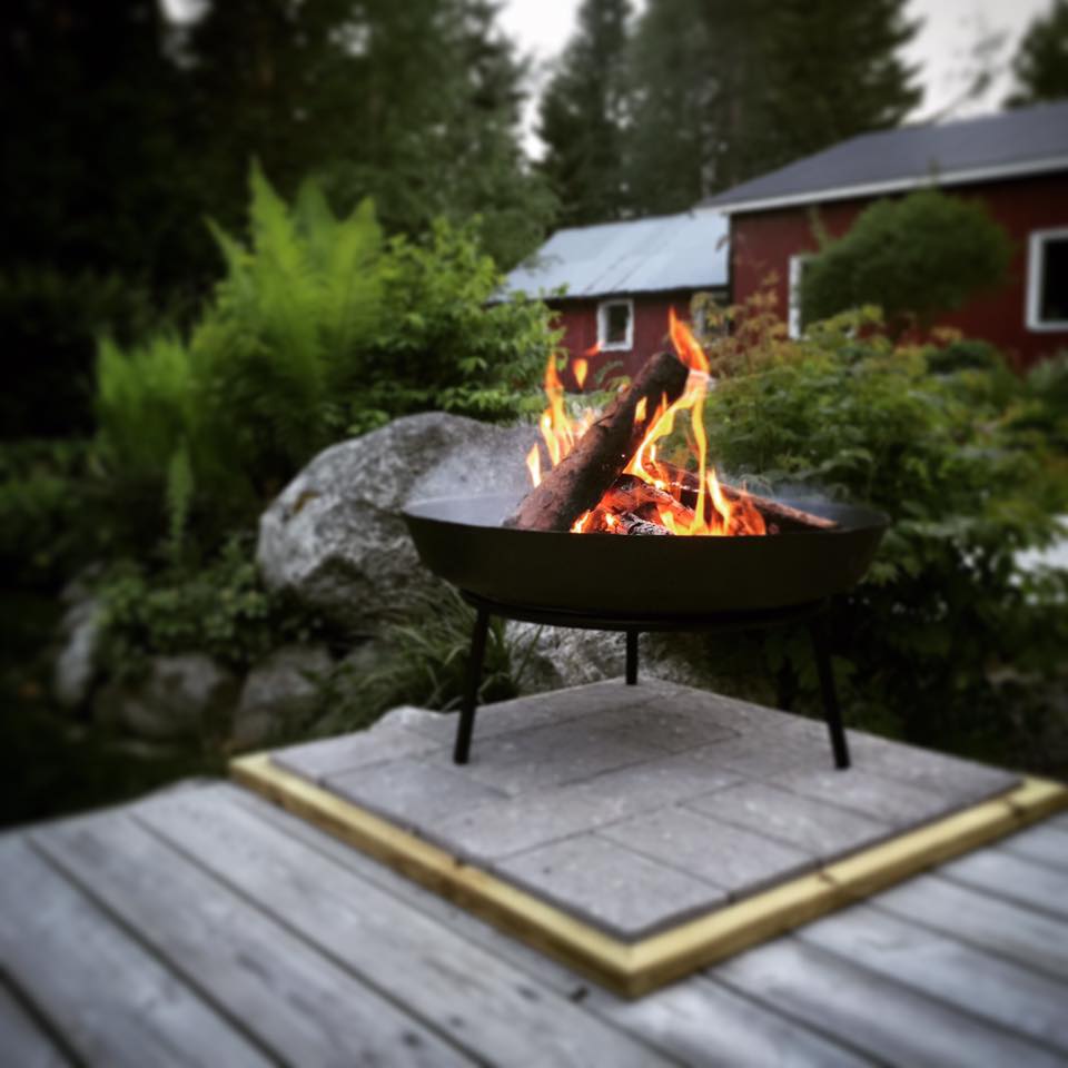 En eldstad med en brinnande stock står på ett trädäck, omgiven av frodig grönska och stenar, med ett rött hus och träd i bakgrunden.