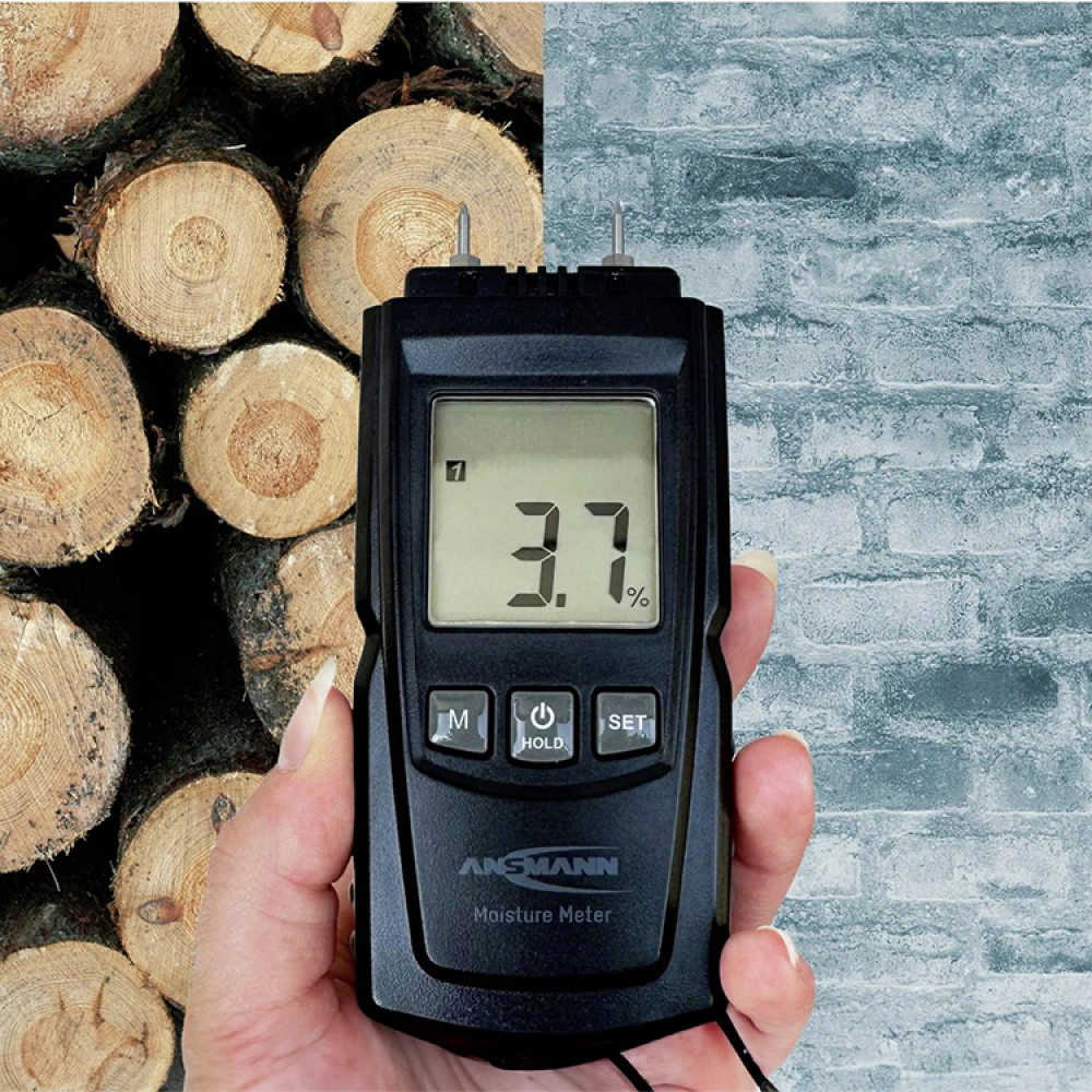 En hand håller i en digital fuktmätare som visar 3,7%, med staplade stockar till vänster och en grå tegelvägg till höger.