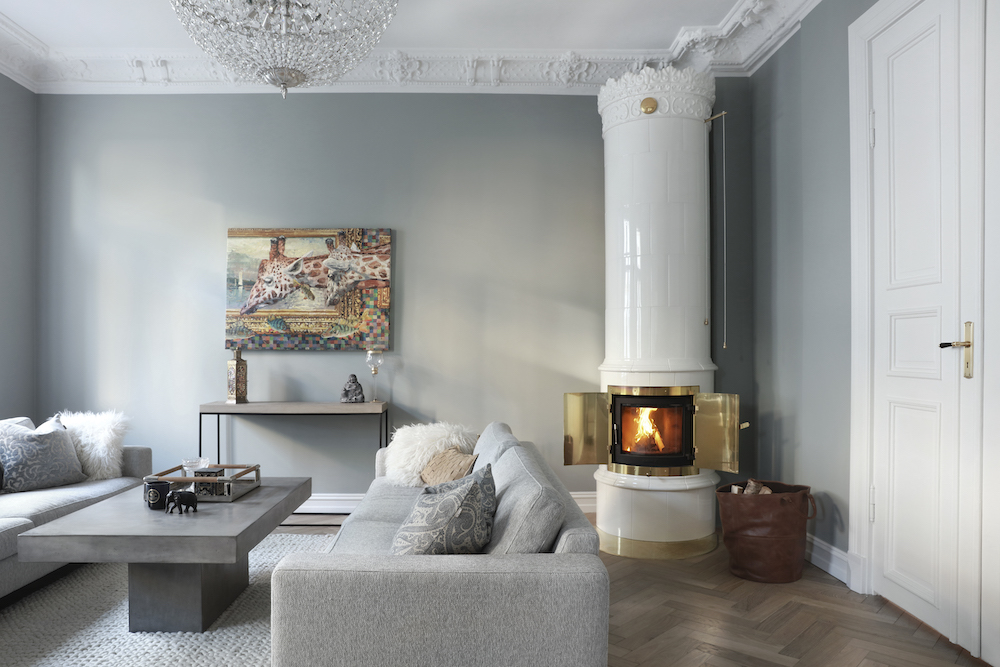 Ett modernt vardagsrum med ljusgrå väggar, en vit utsmyckad öppen spis med en brasa som brinner, två ljusgrå soffor, ett soffbord i betong och en tavla ovanför ett konsolbord. Rummet har dekorativa lister och en ljuskrona.