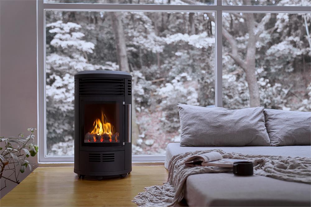 En mysig inomhusmiljö med en modern svart braskamin som brinner starkt bredvid en soffa med kuddar och en filt, en kopp och en bok på ett träbord och stora fönster som visar snöiga träd utanför.