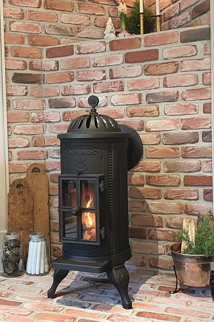 Sortiment 26 En svart vedspis i gjutjärn med en synlig eld inuti står mot en röd tegelvägg. I närheten finns skärbrädor, glasburkar och en krukväxt som skapar en mysig, rustik atmosfär.