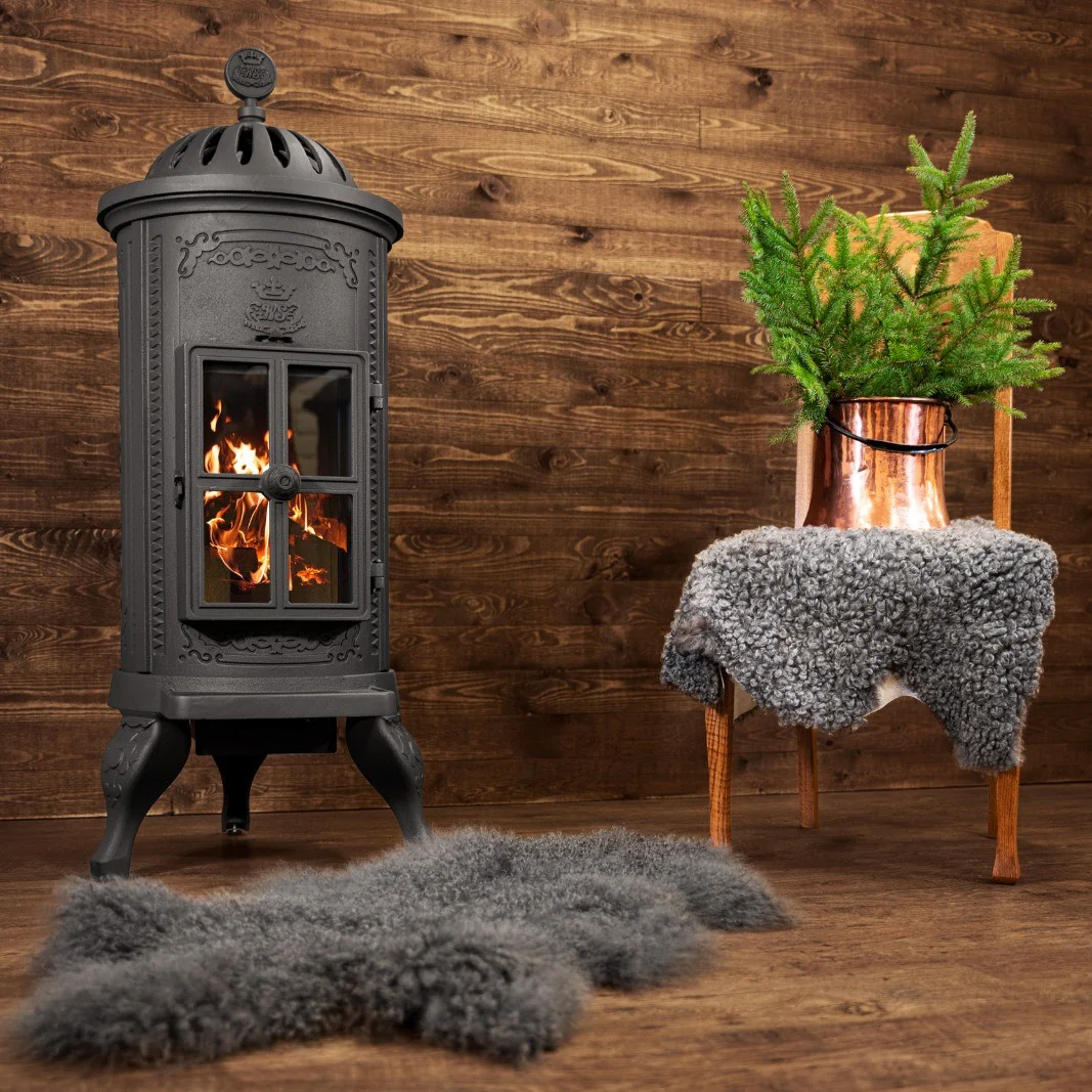 En svart vintage vedeldad kamin med synlig eld står på ett trägolv bredvid en trästol draperad med ett grått fårskinn och en kopparkanna med gröna tallgrenar, mot en träväggsbakgrund.