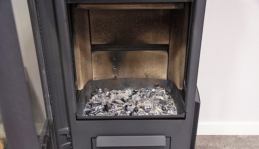 Varför är min kamin svår att tända? 5 vanliga orsaker och enkla lösningar 5 Närbild av en vedspis med metallinredning, med aska och delvis förbrända vedrester i botten. Spisluckan är öppen och visar eldstaden och de sotiga ytorna på insidan.