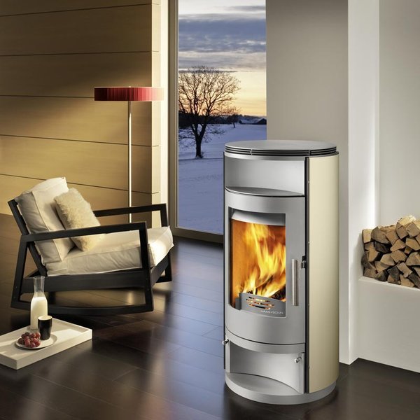 Haas + Sohn: En guide till varumärket 4 Ett modernt vardagsrum med en elegant braskamin, en trave ved, en svart fåtölj med vit kudde, en röd golvlampa och ett stort fönster som visar ett snöigt landskap i solnedgången.