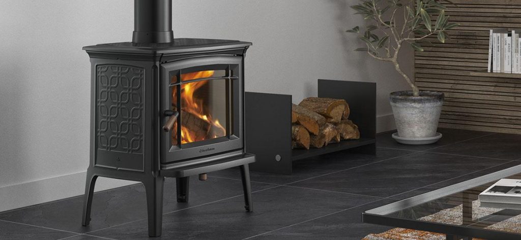 En svart vedeldad kamin med synlig eld inuti står på ett mörkt klinkergolv. Bakom den förvaras staplade vedträn bredvid en krukväxt i ett modernt vardagsrum.