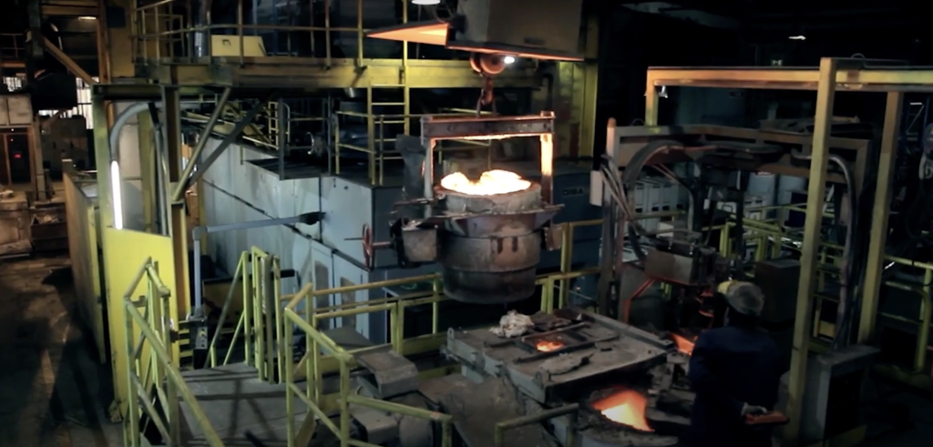 En arbetare övervakar smält metall som hälls från en stor behållare i formar i ett industrigjuteri med gula räcken och tunga maskiner som omger arbetsområdet.
