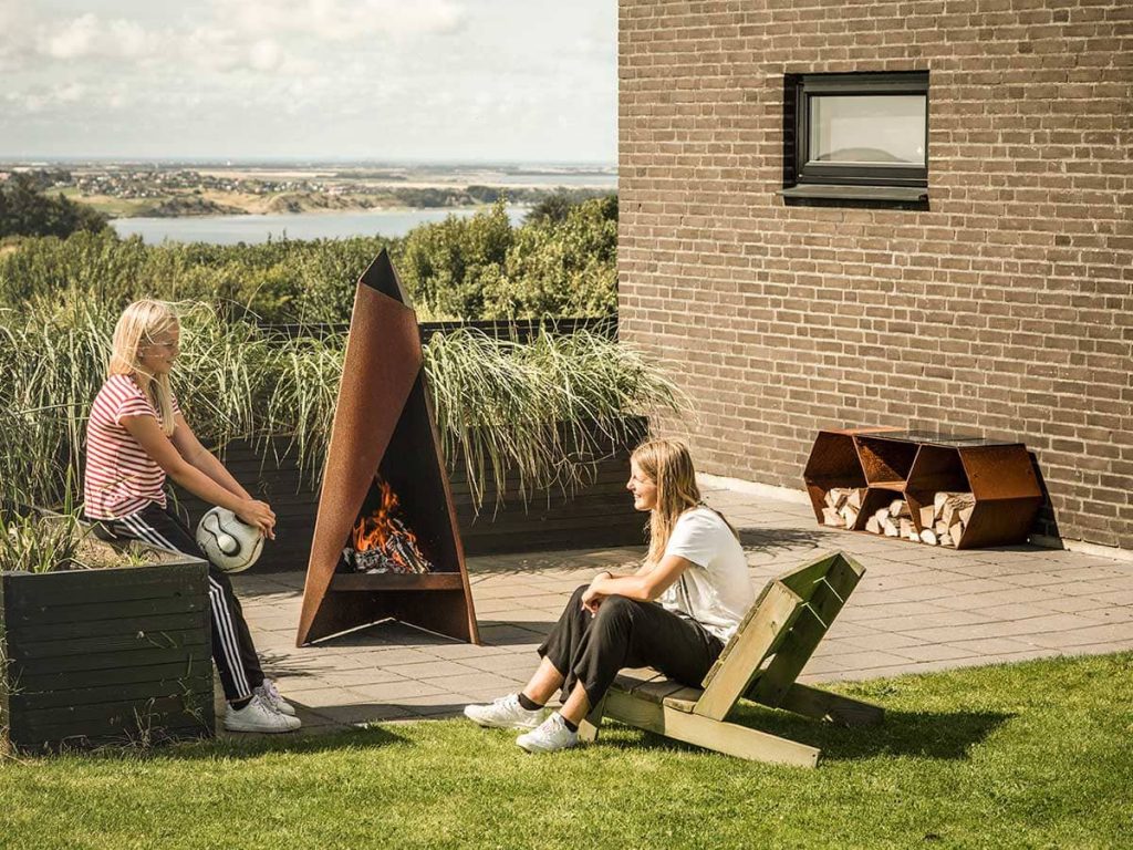 Två personer sitter utomhus vid en modern utomhusspis med triangulär design. Den ena sitter på en planteringskant, den andra på en trästol, med staplad ved och grönska i närheten och en naturskön utsikt i bakgrunden.