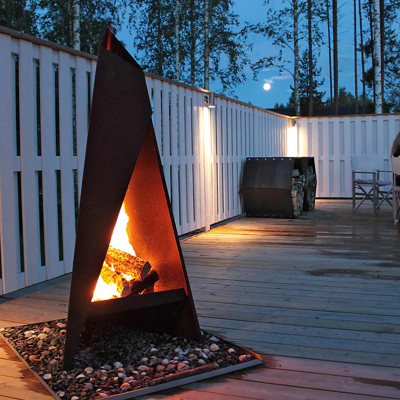 Säker Eldning: Den kompletta guiden (Brandskydd & Regler) 4 En modern triangulär utomhusspis brinner starkt på ett trädäck omgivet av ett vitt staket i skymningen, med träd och månen synlig i bakgrunden.