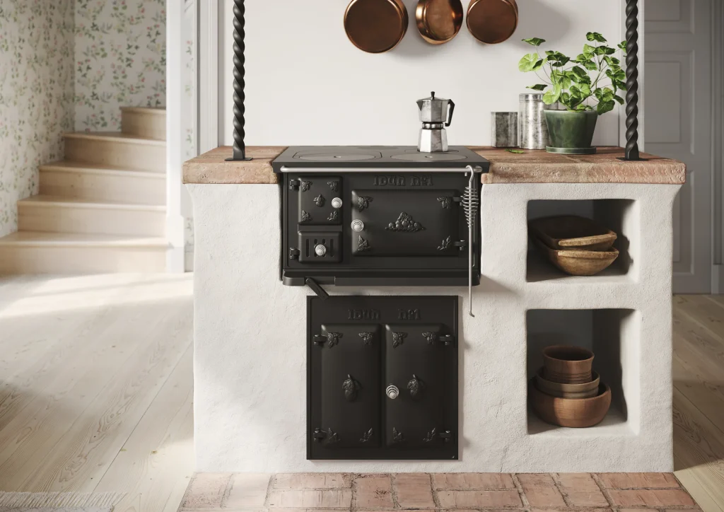 En svart vedeldad vintage-spis är inbyggd i en vit köksö med bänkskiva i terrakotta. Kopparpannor hänger ovanför och på hyllor finns skålar och en krukväxt. Trappor och blommiga tapeter finns i bakgrunden.