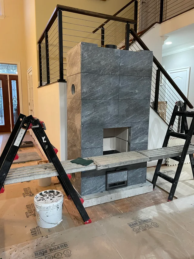 En delvis uppbyggd öppen spis med grå stenplattor står nära en trappa med modernt räcke. Två stegar och en träplanka bildar en provisorisk byggnadsställning. Golvet är täckt av skyddspapper och byggmaterial.