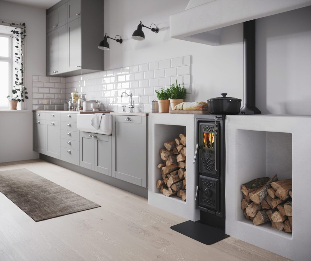 Modernt kök med ljusgrå skåp, vita kakelplattor, bänkskiva i trä, diskho och en svart vedeldad spis med stockar prydligt staplade i inbyggda hyllor på båda sidor.