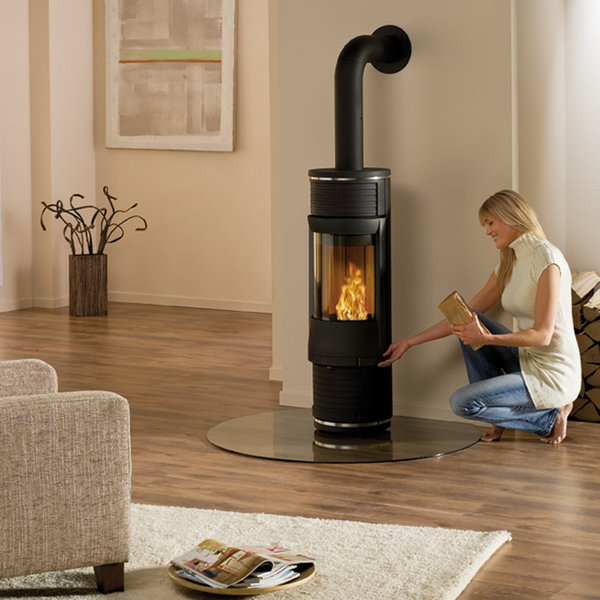 En kvinna knäböjer bredvid en modern svart vedeldad spis i ett mysigt vardagsrum och lägger en stock i elden. Rummet har trägolv, en beige soffa och väggkonst.