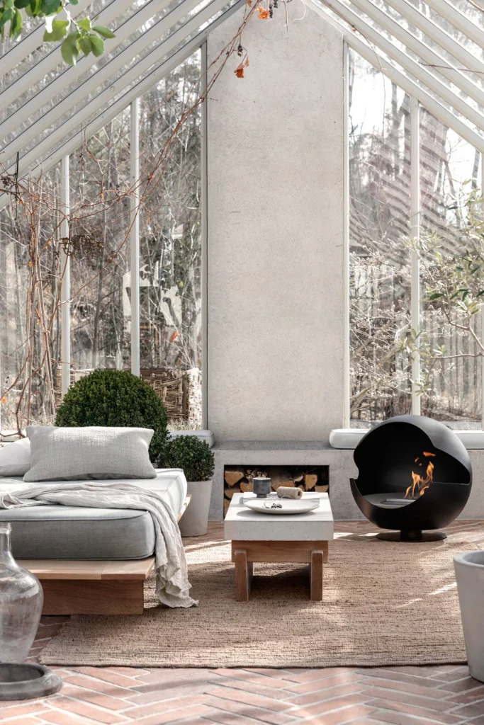 Ett modernt uterum med glasväggar, en stoppad träbänk, ett litet soffbord, en svart öppen spis med synlig låga och krukväxter på en vävd matta, med naturligt ljusinsläpp.