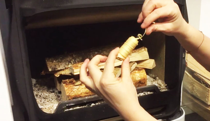 Händer håller i en tändare framför staplad ved i en kamin och förbereder sig för att tända en eld. Aska syns i botten av kaminen.