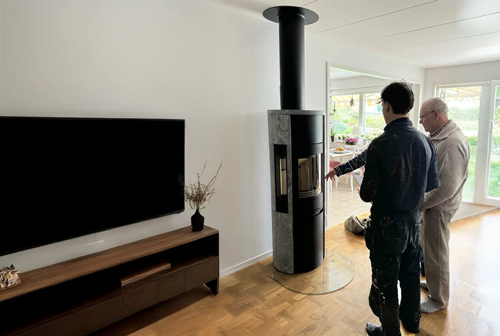 Två personer står i ett vardagsrum bredvid en modern svart vedspis. Den ena pekar på spisen medan den andra iakttar. En TV är monterad på den motsatta väggen ovanför ett träskåp.