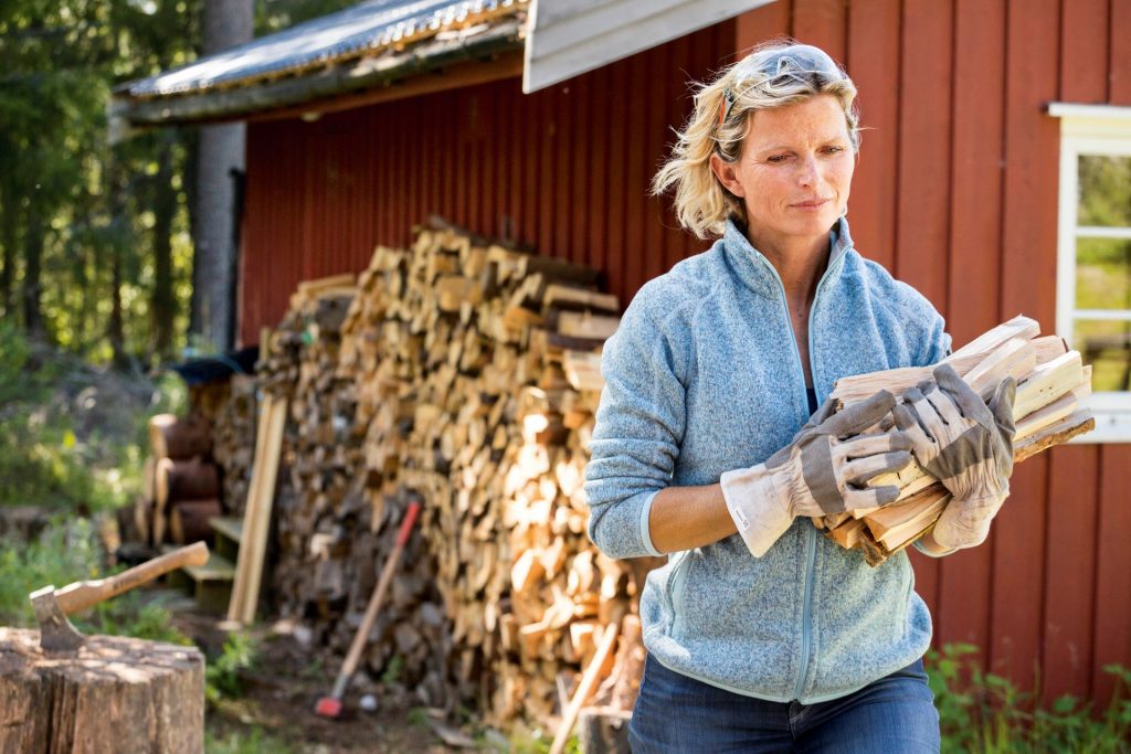 En kvinna iförd handskar och blå fleecejacka bär ved framför en stor vedhög som står staplad mot en röd träbyggnad i en lantlig utomhusmiljö.