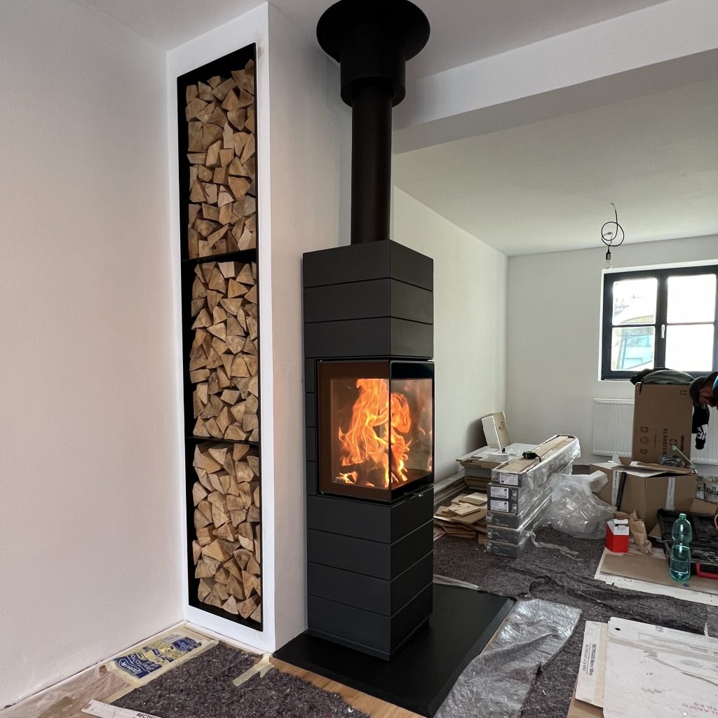 En modern svart braskamin med glasdörr och synliga lågor står bredvid ett högt, inbyggt vedförråd med prydligt staplade vedträn i ett rum som är under renovering.