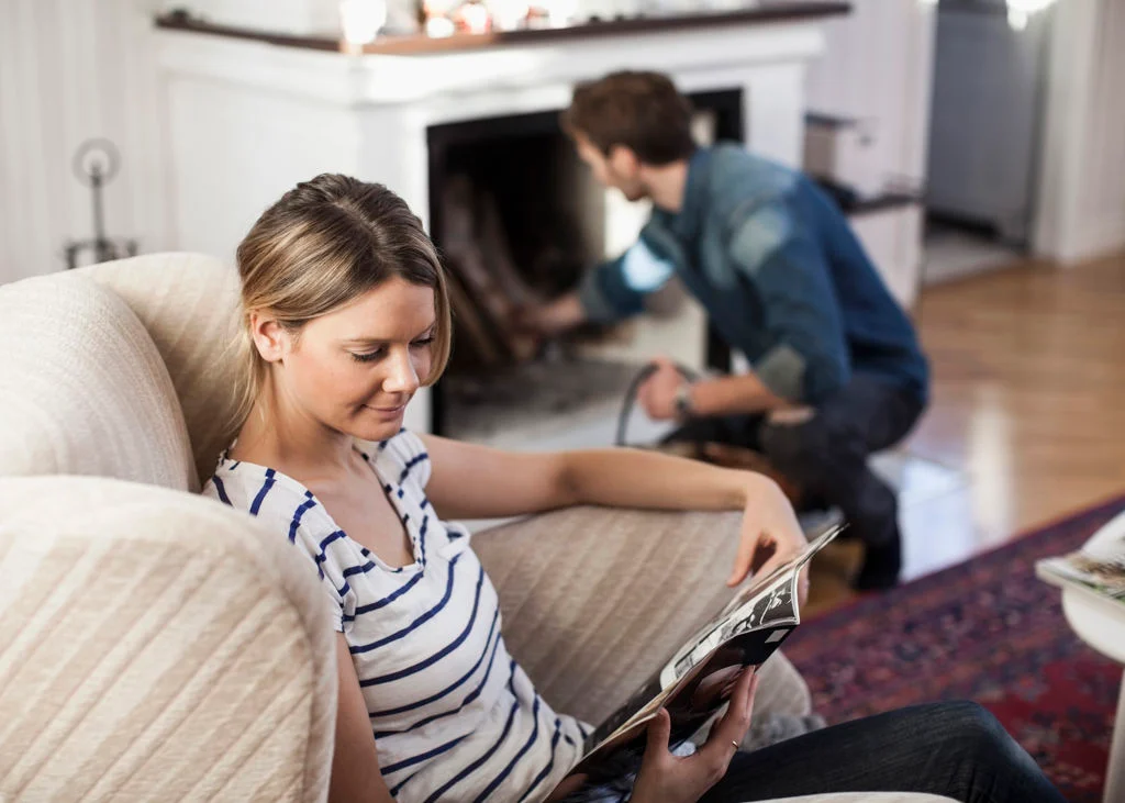 En kvinna sitter i en fåtölj och läser en tidning medan en man står på knä vid en öppen spis i bakgrunden och sköter om den. Rummet är mysigt inrett med bland annat en vit stol och en mönstrad matta.