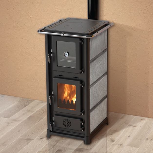 En svart vedeldad kamin med glasdörr som visar en eld inuti står mot en beige vägg på ett ljust trägolv. Kaminen har en topp av metall och sidor av panel.