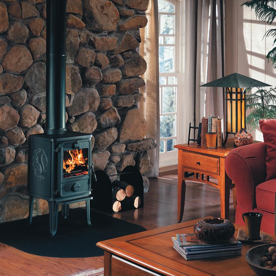 Ett mysigt vardagsrum med en brinnande vedspis framför en stenvägg, vedstaplar i närheten, trämöbler, en röd soffa och solljus som flödar in genom stora fönster med beige gardiner.