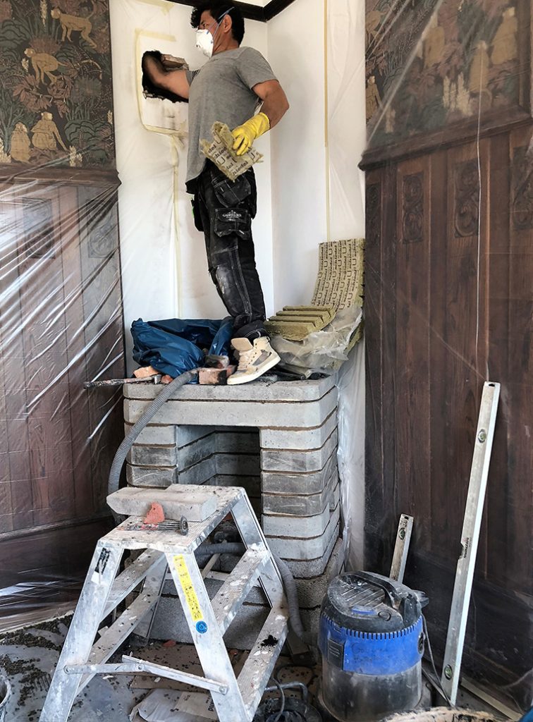 Gabriel Kakelugnar: En guide till svenska klassiker 5 En person iförd dammask och handskar står på en stapel tegelstenar och sticker in armen i ett hål i väggen ovanför en övertäckt öppen spis, omgiven av verktyg, en stege och skyddande plastskynken.