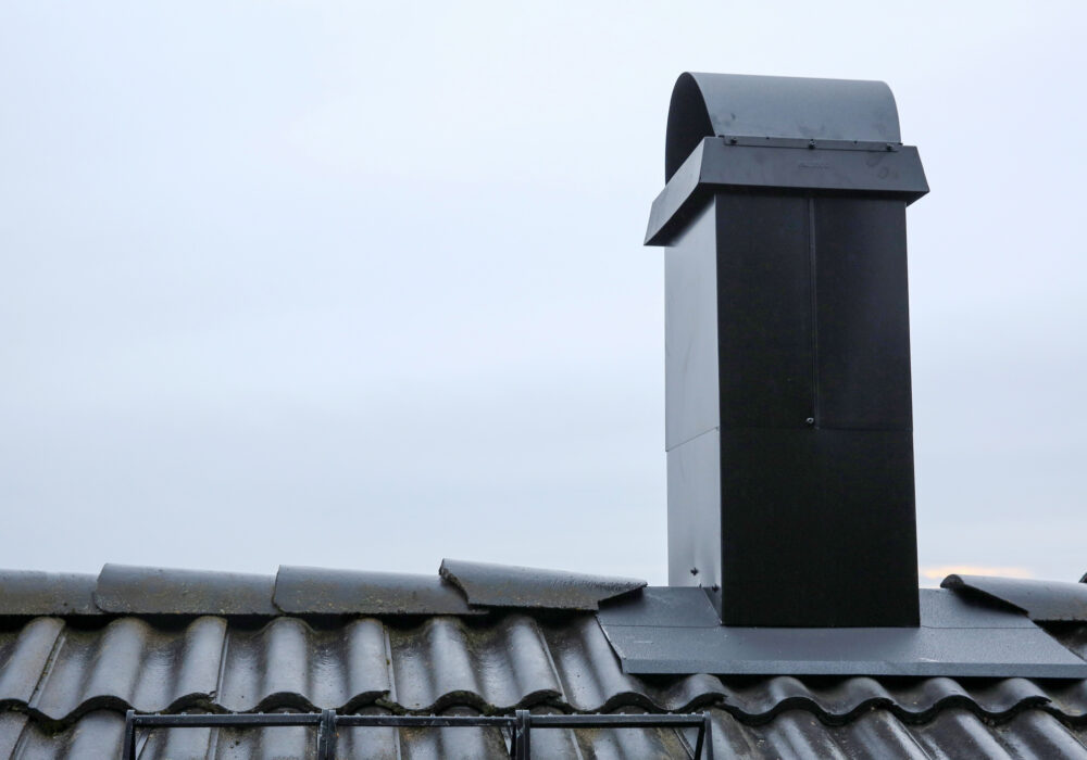 En svart metallskorsten med svängd huv monteras på ett grått tegeltak under en mulen himmel.