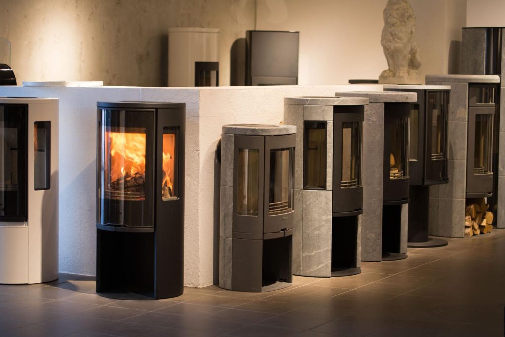 I ett utställningsrum visas en rad moderna braskaminer i olika färger och utföranden. En svart kamin till vänster har en synlig eld som brinner inuti. Bakgrunden består av en vit vägg och ett stengolv.
