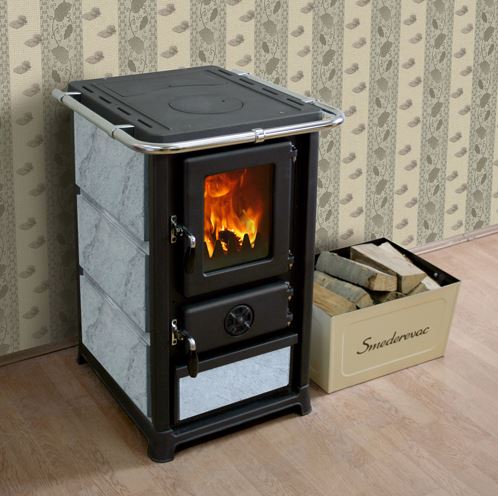 MBS Kaminer & Vedspisar: Den kompletta guiden till ett prisvärt val 3 En liten svartgrå vedspis med en glasdörr som visar en eld inuti, står på ett trägolv bredvid en låda med ved märkt "Smederevac". Mönstrad tapet finns i bakgrunden.