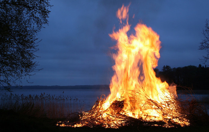 En stor brasa brinner starkt nära en sjöstrand under en mörk, molnig kvällshimmel, med lågor som stiger högt och omges av torra grenar och träd i bakgrunden.