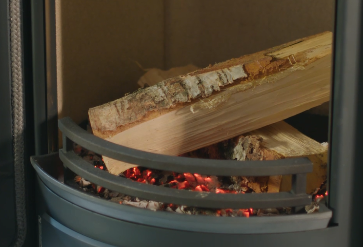 Två vedträn vilar på glödande röd glöd i en metallspis, med metallstänger framför och en brun bakgrund.