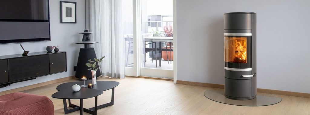 Modernt vardagsrum med ljust trägolv, en rund svart braskamin med en eld inuti, en glashärd, ett svart soffbord med ljus och växter samt stora fönster som leder till en balkong.