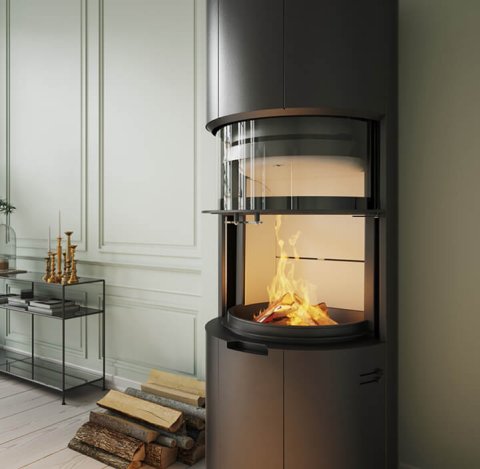 En modern svart cylindrisk öppen spis med glasfront visar en brinnande eld. Staplade vedträn och ljusstakar i guld står på ett konsolbord i närheten i ett ljust, elegant rum med dekorativa väggpaneler.