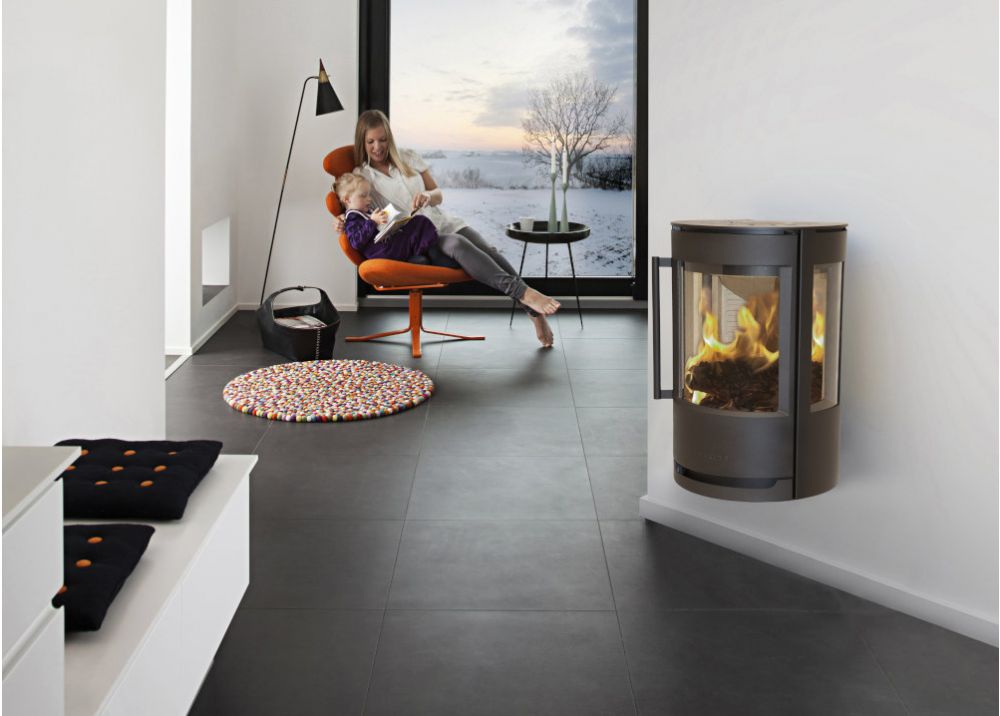 En kvinna sitter i en orange stol och läser för ett barn i ett modernt rum med öppen spis. Ett stort fönster visar ett snöigt landskap och ett kala träd utanför. Rummet är minimalt med en golvlampa, kuddar och mörkt klinkergolv.