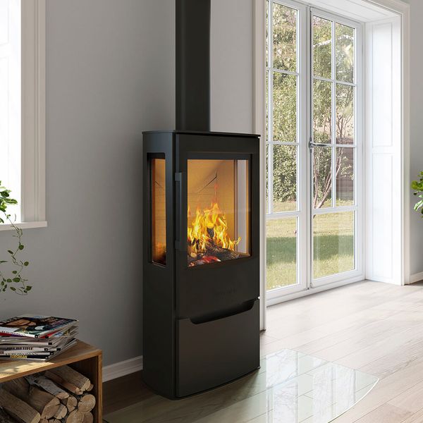 En modern svart vedeldad kamin med synlig eld inuti står nära ett stort fönster i ett ljust rum med trägolv och en trave ved i närheten.