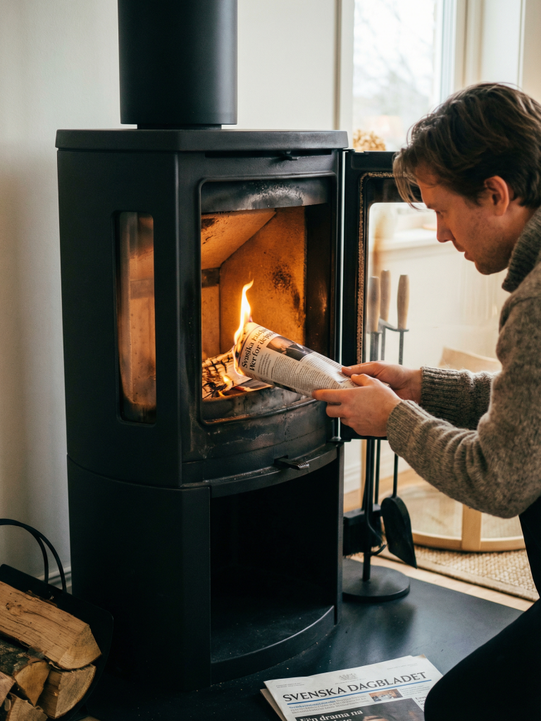 Dåligt drag i skorstenen? (Orsaker & Lösningar som fungerar) 2 En person står på knä vid en svart vedeldad kamin och tänder en brasa med en hoprullad tidning. Luckan till kaminen är öppen och ved ligger staplad i närheten. En svensk dagstidning ligger på golvet.