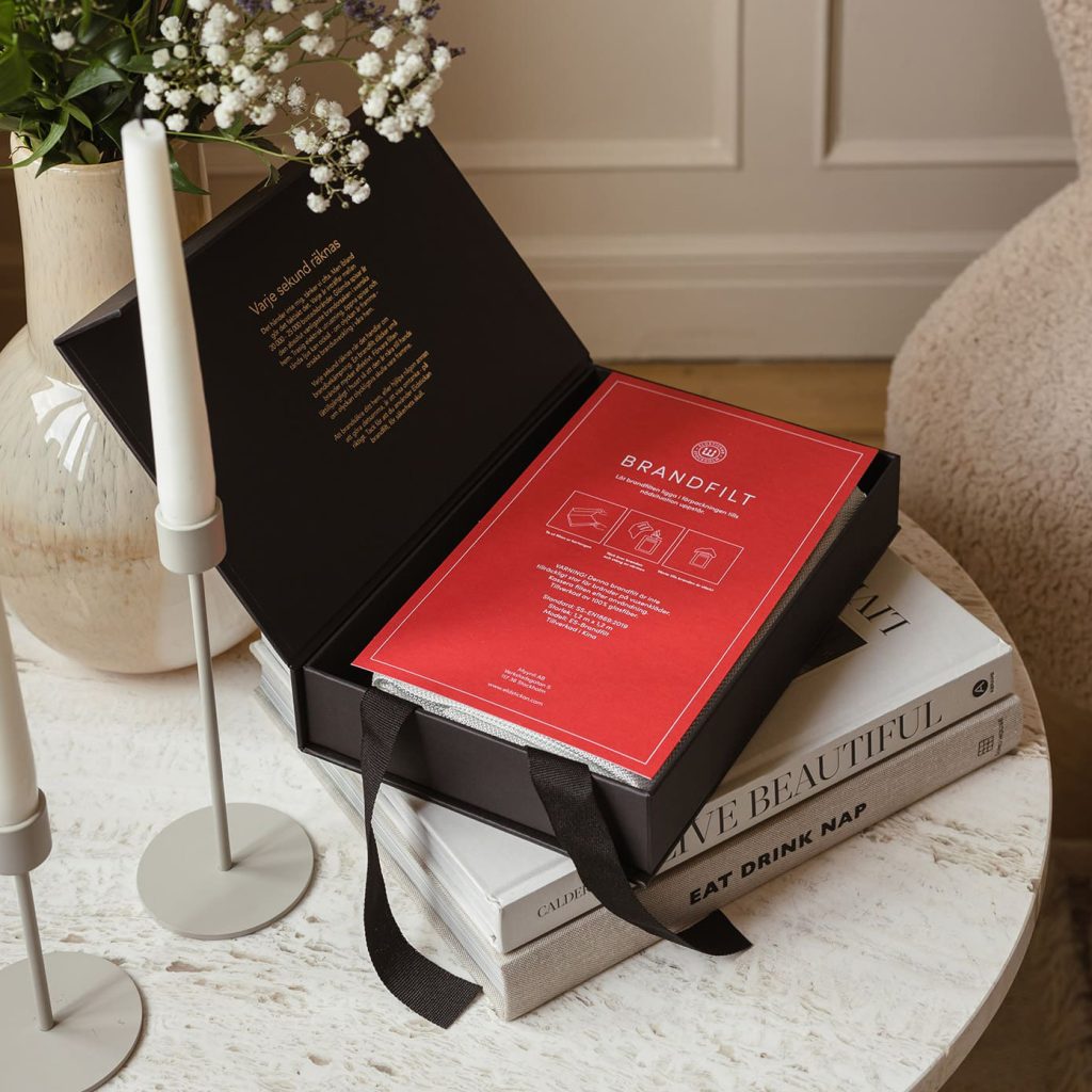 Brandfilt & Brandsläckare: Bäst i Test (Guide för hemmet) 4 En svart presentförpackning med ett rött BRANDFILT-häfte inuti ligger öppen på ett runt bord, bredvid staplade böcker, levande ljus och en vas med blommor i ett mysigt, stilfullt rum.