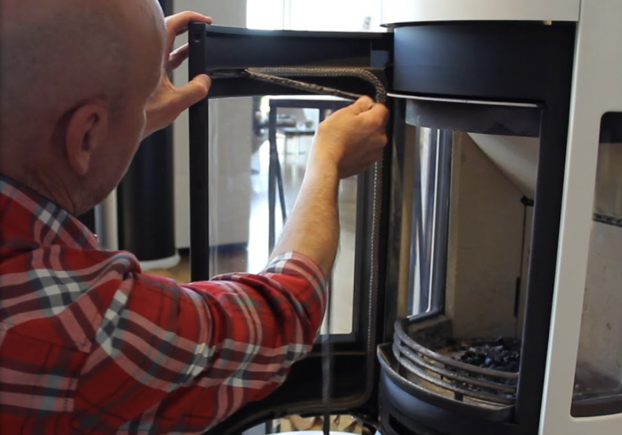 En person i rödrutig skjorta byter ut tätningen på glasdörren till en insats i en öppen spis, med dörren öppen och insidan av den öppna spisen synlig.