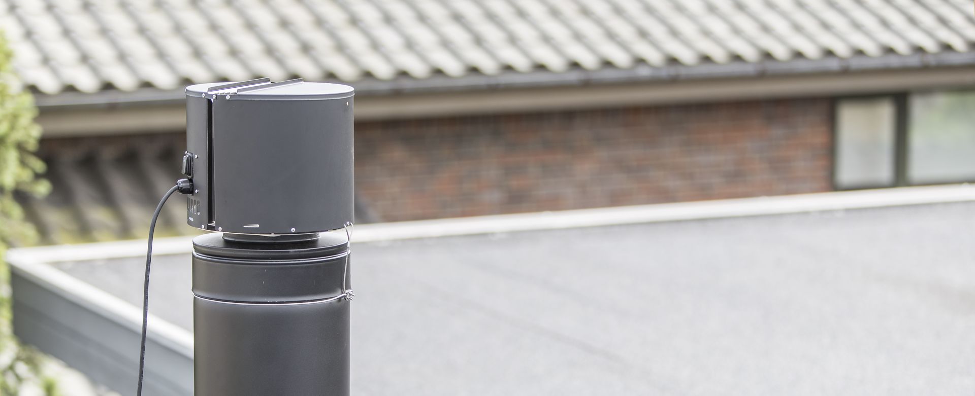 Närbild av en svart cylindrisk enhet monterad ovanpå ett runt rör eller en skorsten, med en strömkabel ansluten. I bakgrunden syns ett suddigt hustak med grå bältros och ett fönster.