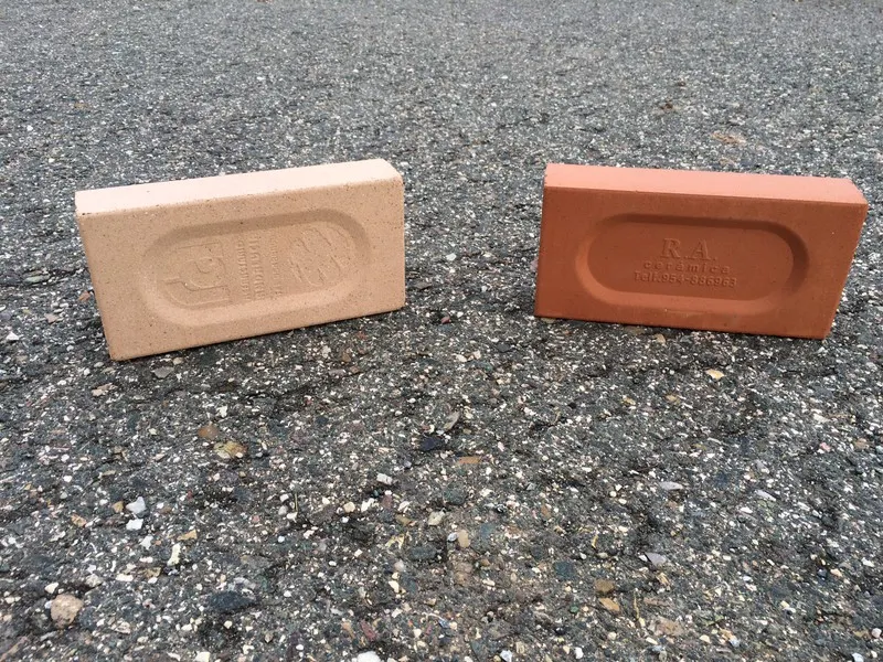Två rektangulära tegelstenar, en ljusbeige och en röd, står upprätt på en grov, grå asfaltsyta. Båda tegelstenarna har indragna markeringar med text, men texten är inte helt läsbar.