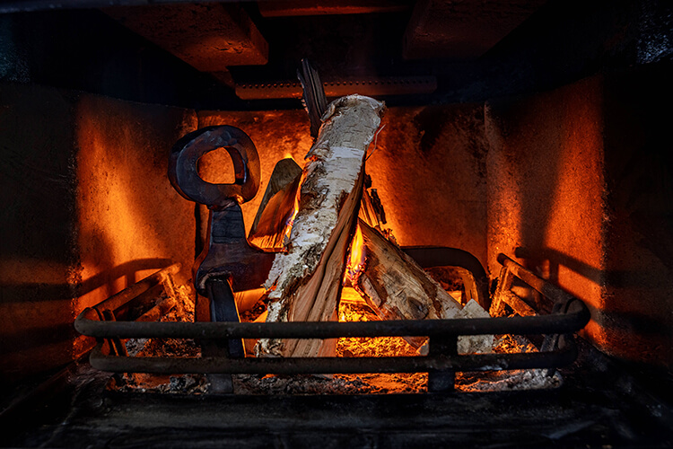 Eldställ & Kaminverktyg: En komplett köpguide (2026) 4 Närbild av en brinnande öppen spis med glödande glöd, vedträn och ett svart brandskydd i metall. Varma orange flammor lyser upp interiören och skapar en mysig och inbjudande atmosfär.