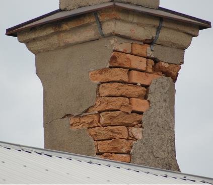 Närbild av en skadad tegelskorsten med stora sprickor och saknad ytterputs, som avslöjar tegelstenarna undertill. Skorstenen står på ett plåttak mot en molnig himmel.