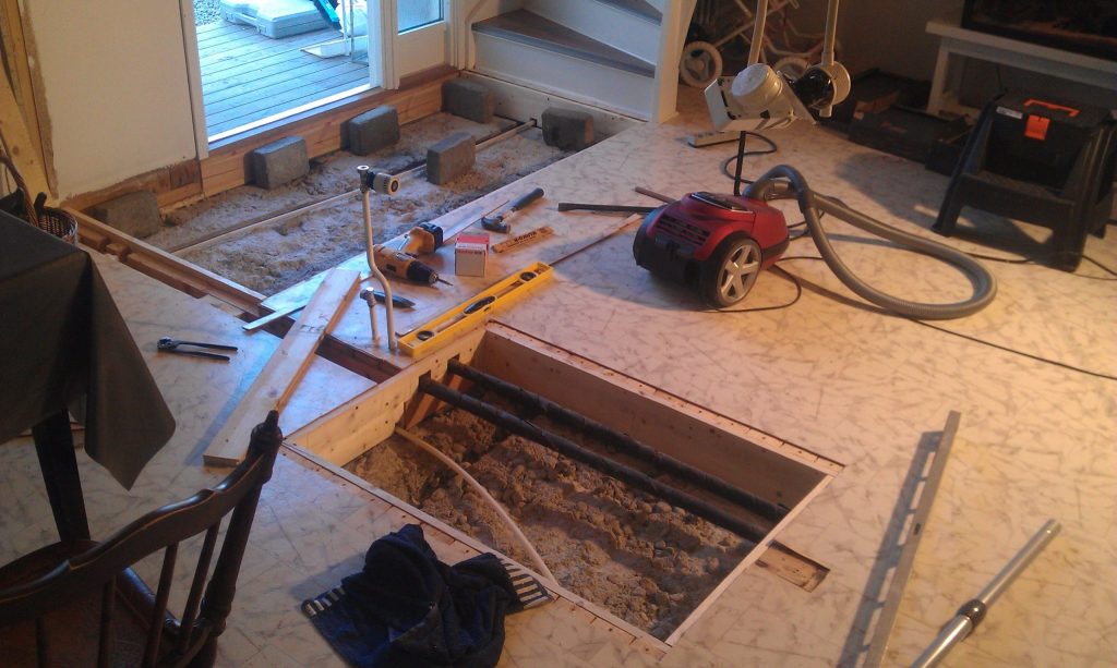 Ett rum under renovering med en del av golvet borttaget, vilket blottlägger rör och sand under. Verktyg, en dammsugare, lampor och byggmaterial ligger utspridda. I bakgrunden syns en skjutdörr i glas och slaggblock.