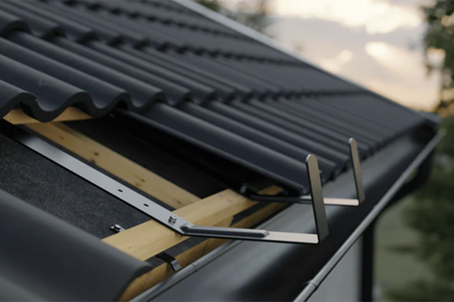 Närbild av ett mörkfärgat plåttak med vågformade paneler och synliga träbjälkar, med ett snörasskydd av metall fäst nära takkanten mot en suddig bakgrund utomhus.