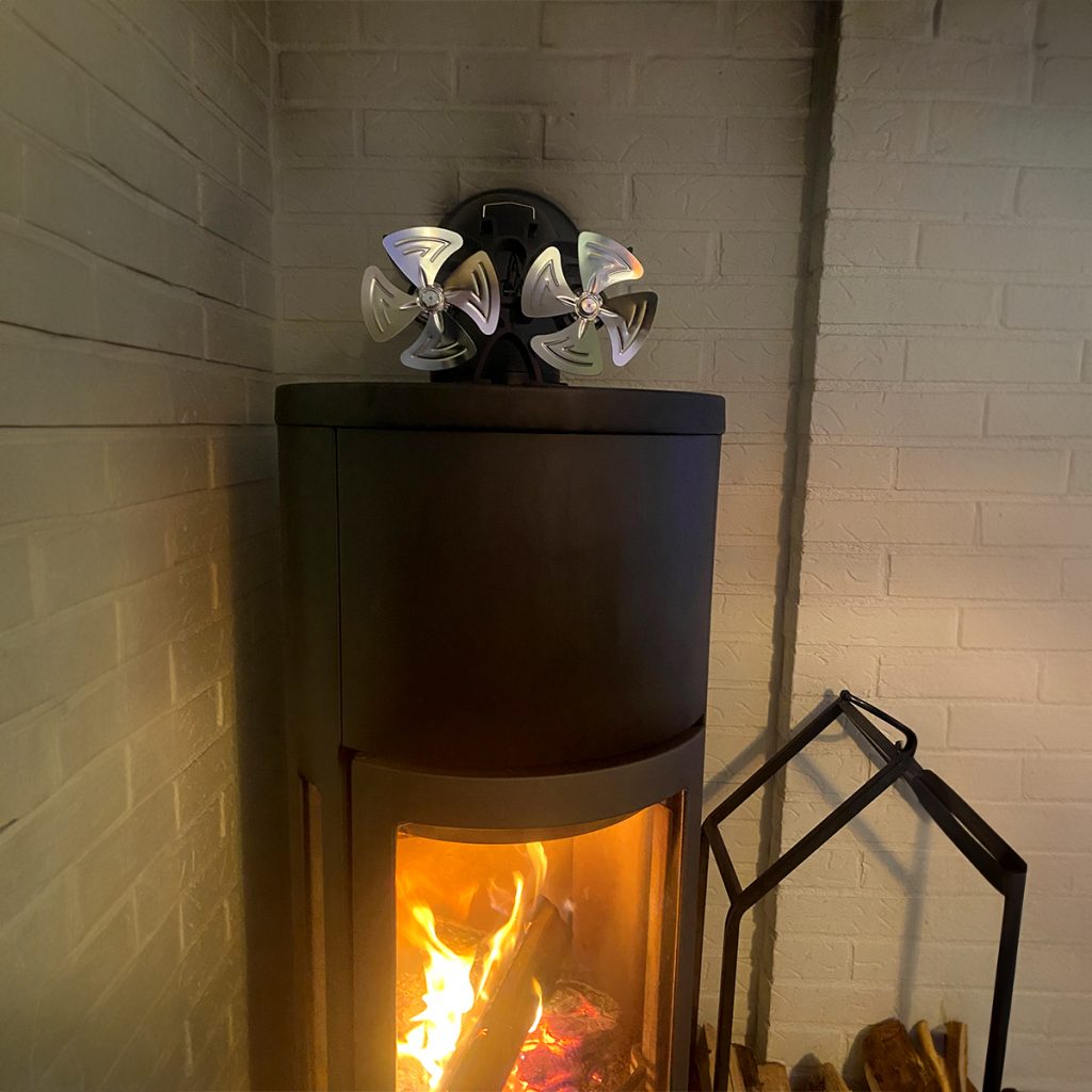 En vedeldad kamin med synlig eld inuti står i ett hörn och toppas av en fläkt i metall. Veden ligger staplad i en vedhållare bredvid kaminen mot en vit tegelvägg.