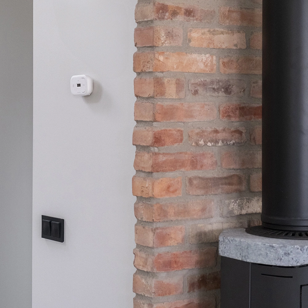 Säker Eldning: Den kompletta guiden (Brandskydd & Regler) 3 Del av innervägg med synligt rött tegel intill en vitmålad vägg. En termostat och två svarta strömbrytare är monterade på den vita väggen och till höger syns en del av en svart vedspis med stentopp.