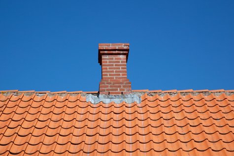 En skorsten i rött tegel, utformad för att förhindra kondens i skorstenen, sträcker sig över ett tak täckt med orange, vågiga keramikplattor, mot en klarblå himmel.