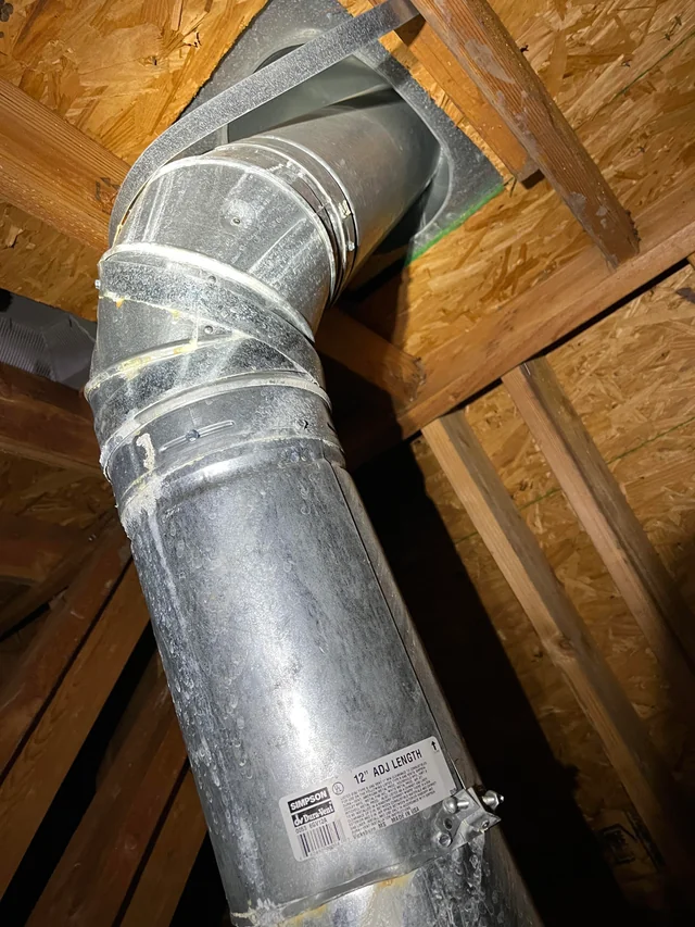 En luftkanal av metall på en vind är fäst med buntband i träbjälkar, med isolering och byggmaterial synliga runtomkring. På en etikett på kanalen står det 12 ADJ. LÄNGD.