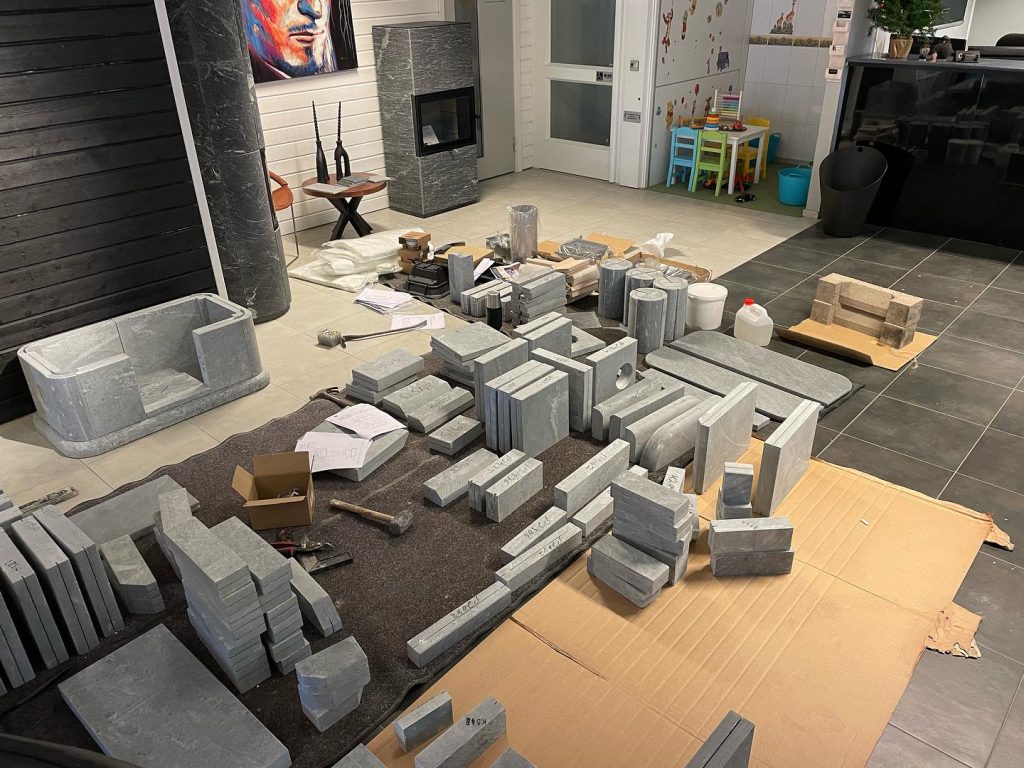 Grå stenblock i olika storlekar är prydligt uppställda på kartong och en matta inomhus, med verktyg och papper utspridda runt omkring. I bakgrunden syns ett lekbord, en spis och ett konstverk.